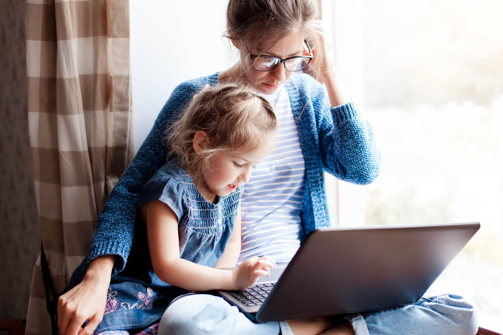 Working Mum works from home office with kid. Mother and daughter wearing blue and looking at a laptop.