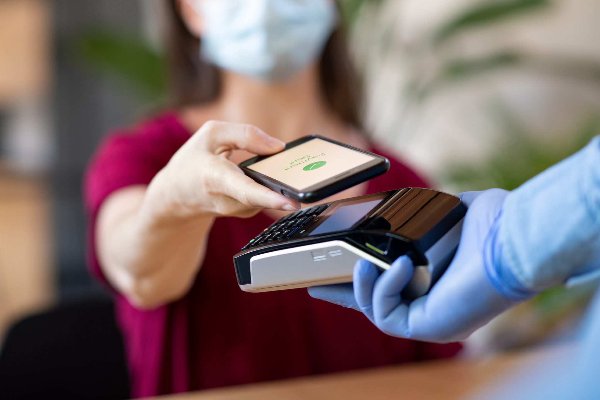Close up hand of customer paying with smartphone. Cashier hand holding credit card reader machine and wearing protective disposable gloves at bar counter, while client holding phone for payment. Woman wearing face mask while paying bill with mobile phone during Covid-19 pandemic.