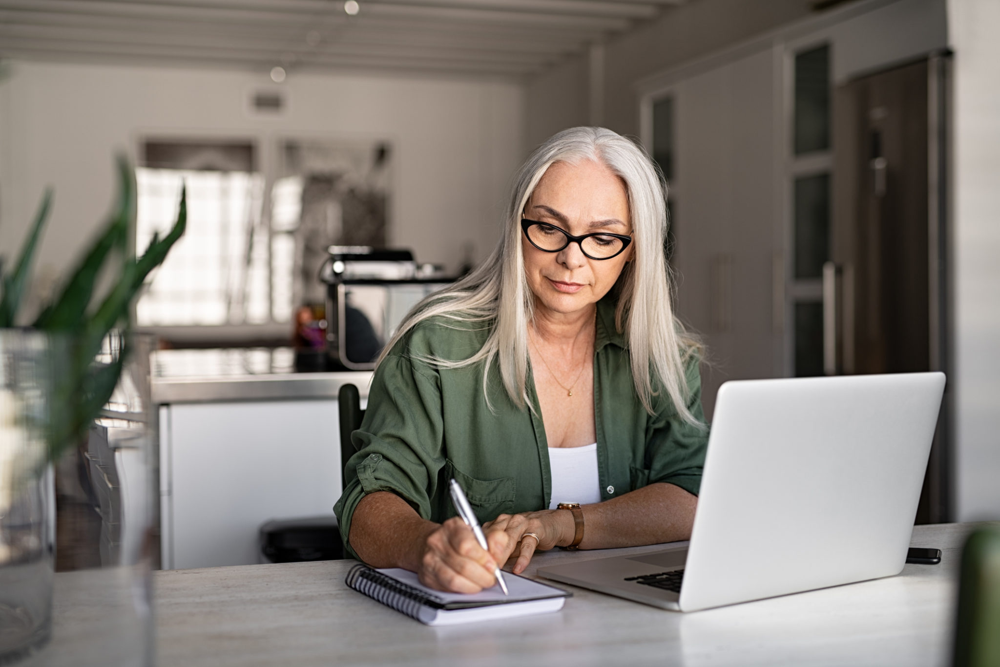 Senior stylish woman taking notes in notebook while using laptop to follow a step by step guide on how to perform an SEO audit on her small business website