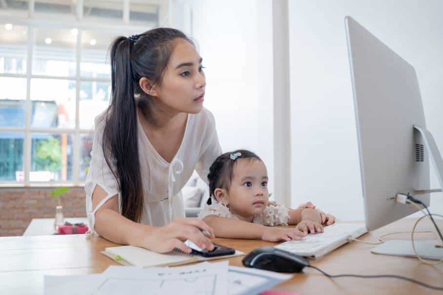Woman working from home on an apple mac computer wearing a white top and long ponytail with her daughter sat on her lap
